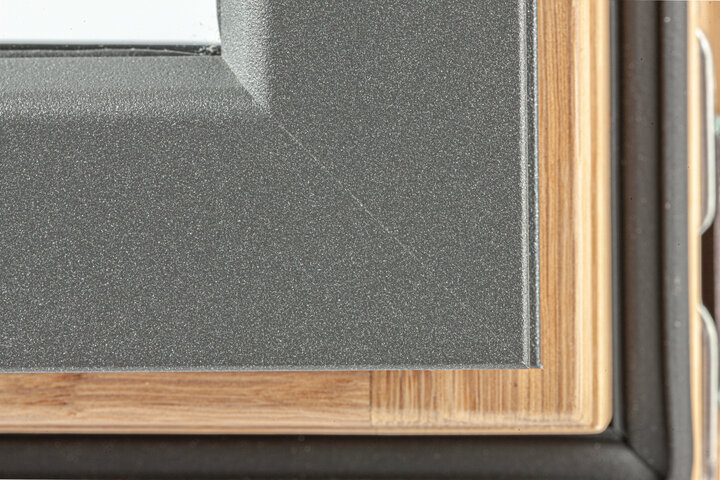 Eine graue Metalloberfläche mit einer subtilen Textur und ein Holzrahmen mit einem hellbraunen Farbton sind in Nahaufnahme zu sehen.