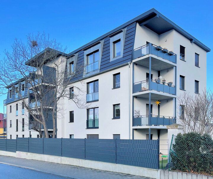Ein modernes Wohnhaus mit moderner Fassade, Balkonen und einem Baum im Vordergrund liegt an einer Straße mit blauem Himmel und einem Zaun.