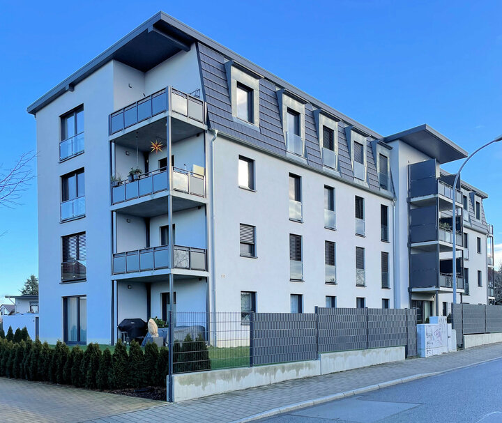 Ein modernes Wohnhaus mit weißen Wänden, grauem Dach und Balkonen steht an einer Straßenecke, begleitet von einem schwarzen Zaun, einer Straßenlaterne und einem klaren blauen Himmel.