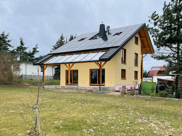 Ein gelbes Haus mit Sonnenkollektoren auf dem Dach, steht auf einem üppigen grünen Rasen, mit einem weißen Schuppen und Bäumen im Hintergrund.