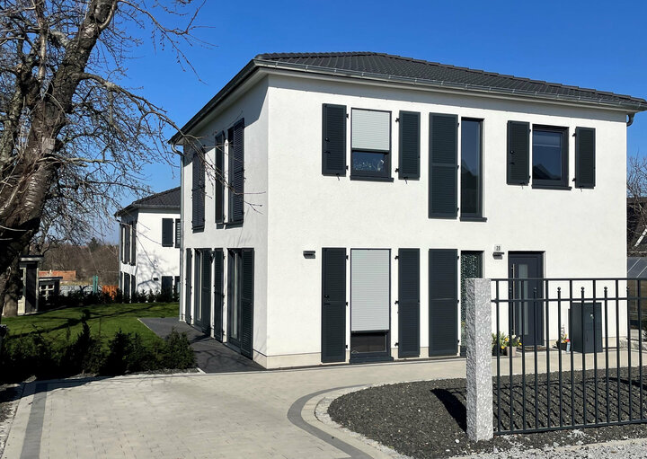 Ein modernes weißes Haus mit schwarzen Fenstern und einem schwarzen Dach befindet sich in einem Vorort, begleitet von einem schwarzen Tor und einem Baum im Vordergrund.