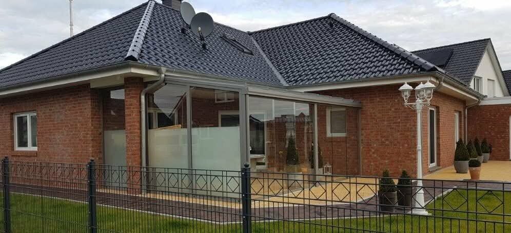 Ein modernes Haus mit schwarzem Dach, Backsteinfassade und großen Glasfenstern ist von einem schwarzen Metallzaun umgeben, daneben stehen ein weißer Laternenpfahl und Topfpflanzen.