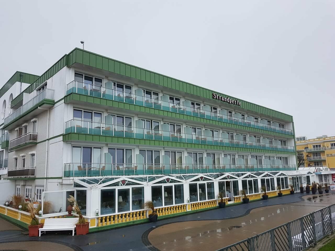 Das Strandperle Hotel, ein weißes Gebäude mit grünen Akzenten, verfügt über Balkone, ein grünes Dach und eine gelbe Markise, mit einem Brunnen und einer Bank im Vordergrund.