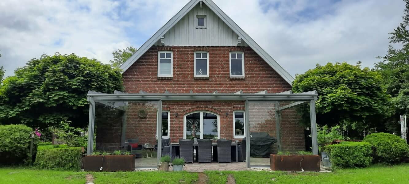 Ein rotes Backsteinhaus mit einem weiß gerahmten Giebeldach und einer verglasten Terrasse mit Tisch und Stühlen ist von üppigem Grün und einem klaren blauen Himmel umgeben.