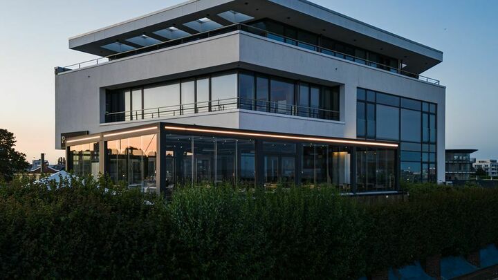 Ein modernes, weißes Gebäude mit Flachdach und großen Fenstern ist von üppigem Grün umgeben, mit einer glasüberdachten Terrasse und Sitzgelegenheiten im Freien.