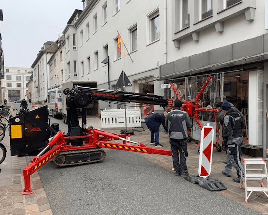 Ein roter Kran steht auf einer Straße in der Stadt, begleitet von Arbeitern und einem weißen Lieferwagen, mit einer rot-weiß gestreiften Absperrung in der Nähe.