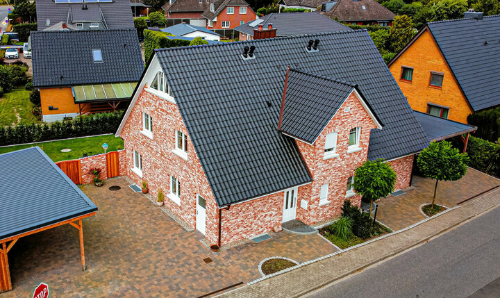 Ein zweistöckiges Backsteinhaus mit schwarzem Dach und Schornstein, umgeben von einer gemauerten Einfahrt und einem kleinen Garten, befindet sich in einem Wohnviertel.