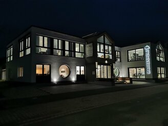 Ein modernes zweistöckiges Gebäude mit großen Fenstern, einer geschwungenen Fassade und einem Schild mit der Aufschrift "MAHRENHOLZ" ist nachts beleuchtet, mit einer Straße und einem Gehweg im Vordergrund.