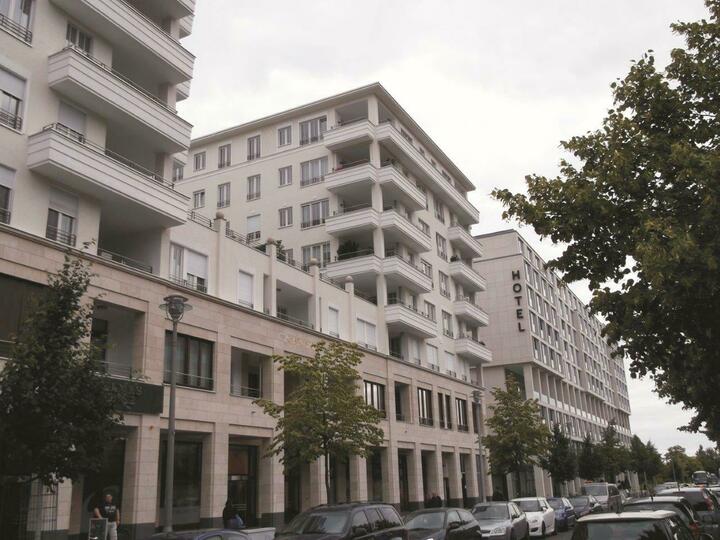 In einer Stadtstraße stehen eine Reihe weißer Wohnhäuser mit Balkonen und ein beiges Gebäude mit einem "HOTEL"-Schild, umgeben von geparkten Autos und Bäumen.
