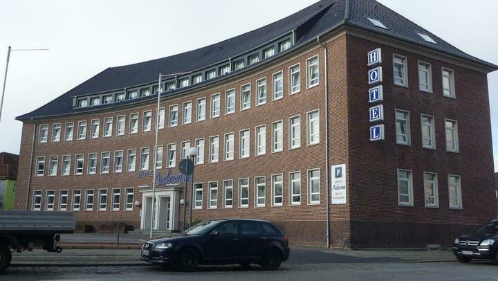 Ein Backsteingebäude mit geschwungener Fassade und "HOTEL"-Schild steht an einer Straßenecke, und in der Nähe parken ein schwarzes Auto und ein weißer Lastwagen.