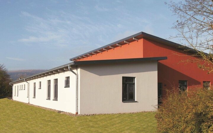 Ein modernes Gebäude mit rot-weißem Äußeren, schwarzem Dach und einem kleinen Garten steht vor einem klaren blauen Himmel und üppigem grünen Rasen.