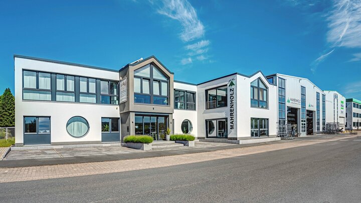 Ein modernes weißes Gebäude mit einem grauen Dach, großen Fenstern und einem Schild für &quot;Mahnenholz&quot; steht an einer Straßenecke, mit einem klaren blauen Himmel und Bäumen im Hintergrund.