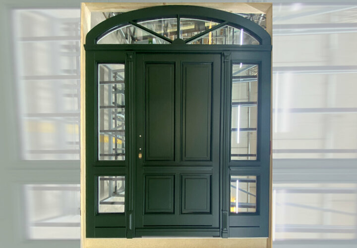 Eine dunkelgrüne Holztür mit einem Oberlicht und zwei Seitenteilen. Das Oberlicht und die Seitenteile sind mit Sprossen ausgeführt.