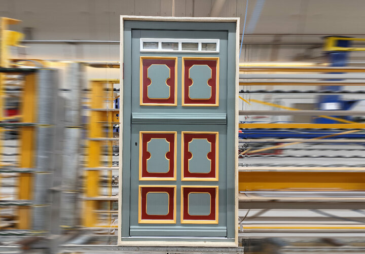 Eine grau-rote Holztür mit gelben Akzenten steht in einer belebten Werkstatt, umgeben von gelben und grauen Maschinen.