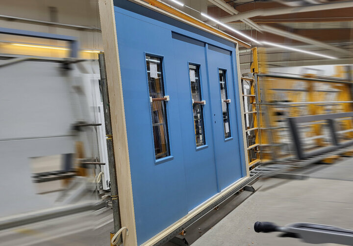 Eine blaue mehrteilige Türe mit drei rechteckigen Lichtausschnitten steht in der Verglasung auf einem Transportband. 
