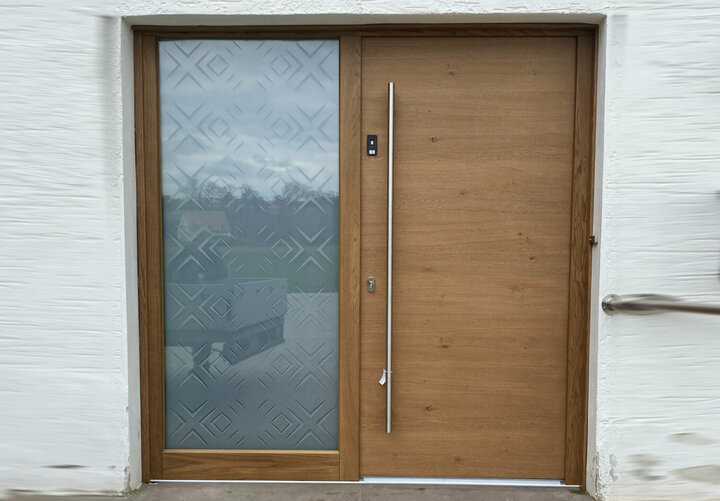 Eine moderne geschlossene Holztür mit einem Seitenteil aus Glas ist in eine weiße Wand eingelassen. Die Tür ist mit einem Stangengriff und einem Fingerprintscanner ausgestattet.