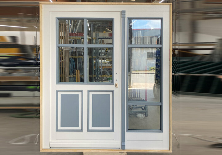 Eine weiße Holztür mit Ganzglasseitenteil und grauen Akzenten, die große Scheibe in der Tür ist mit einem Sprossenkreuz versehen. unter der Glasscheibe befinden sich zwei quadratische Flächen in grauer Farbe.