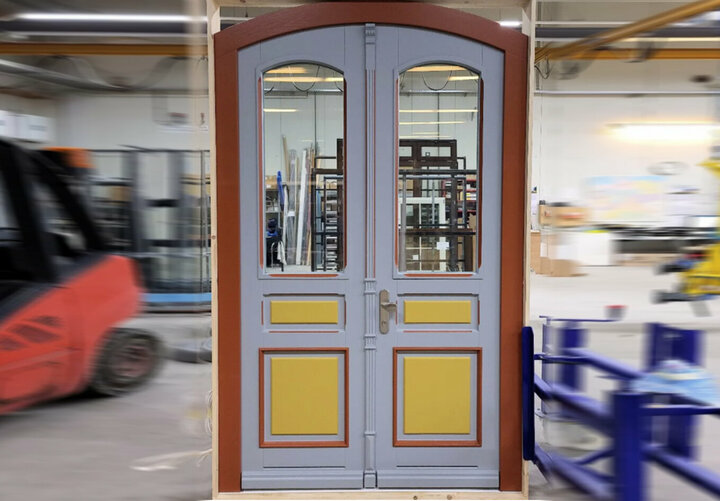 Eine graue Tür mit gelben Akzenten und rotem Rahmen steht in einer belebten Werkstatt, spiegelt die umliegende Aktivität wider und steht im Kontrast zu den weißen Wänden.