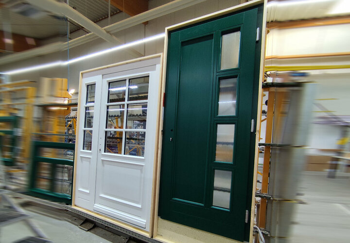 Eine weiße verglaste Tür und eine dunkle Tür mit vier quadratischen Glasausschnitten  sind in einer Halle auf dem Transportband abgestellt.