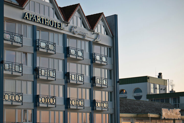 Ein Aparthotel am Strand spiegelt sich im Wasser. Viele Strandkörbe und ein blauer Himmel sind auf dem Bild zu sehen.