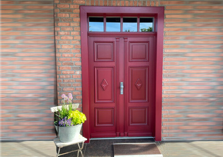Eine rote Doppeltür Oberlicht ist in eine rote Backsteinmauer eingelassen, begleitet von einem weißen Stuhl und einer Topfpflanze.