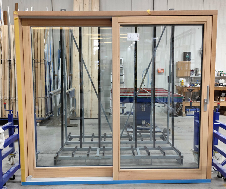 Eine große Schiebetür mit einem hölzernen Rahmen befindet sich in einer geschäftigen Werkstatt, in der eine blaue Werkbank und ein Stapel Holzbretter zu sehen sind.