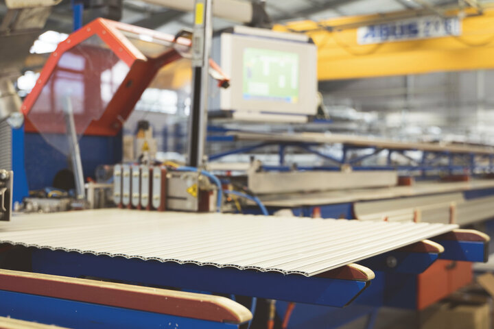 Eine blau-rote Maschine mit einem rot-weiß gestreiften Förderband transportiert Rollladenpanzer in einer belebten Fabrik.