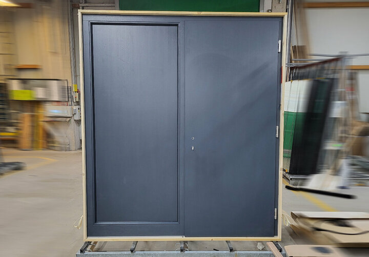 Eine große, dunkelgraue Doppeltür mit silbernem Griff und Schloss steht auf einer Holzpalette in einem belebten Lagerhaus, umgeben von Werkzeugen und Geräten.