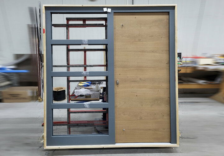 Eine Holztür mit grauem Rahmen und eine Seitenteil geben den Blick auf einen  Arbeitsbereich in einer Lagerhalle frei.
