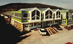 Ein älteres Gebäude mit grün-weißem Äußeren, großen Fenstern und einem Glasdach steht an einer Straßenecke, vor dem ein rotes Auto parkt und im Hintergrund ein Hügel zu sehen ist.