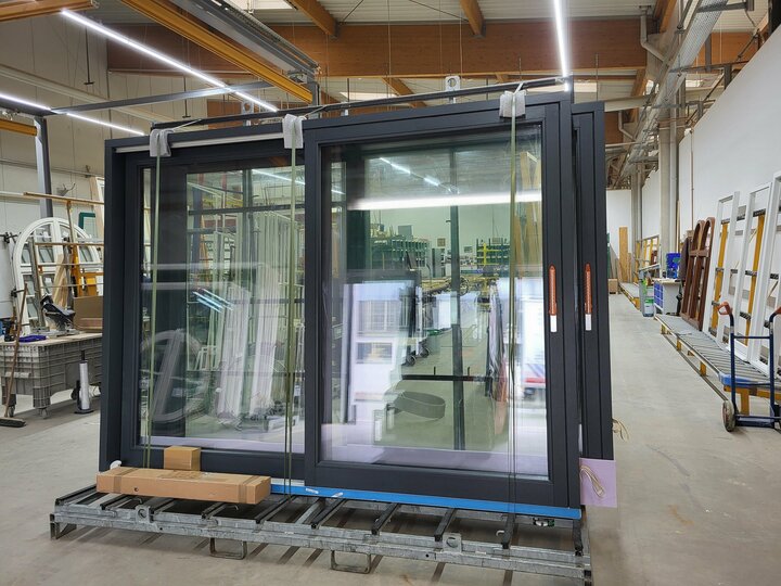 Eine große schwarze Glasschiebetür, die mit Gurten gesichert ist, steht in einem geräumigen Lagerhaus, begleitet von einem blauen Wagen und einer Holzpalette.