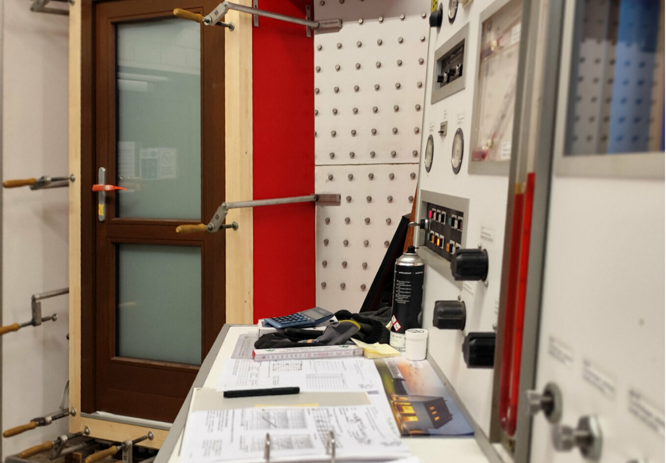 Ein weißes Bedienfeld mit roten und schwarzen Tasten und Knöpfen, ein schwarzer Taschenrechner und ein rot-schwarzer Kanister befinden sich in einer Werkstatt mit roter Tür und weißer Wand. Hier handelt es sich um einen RAL-Prüfstand.