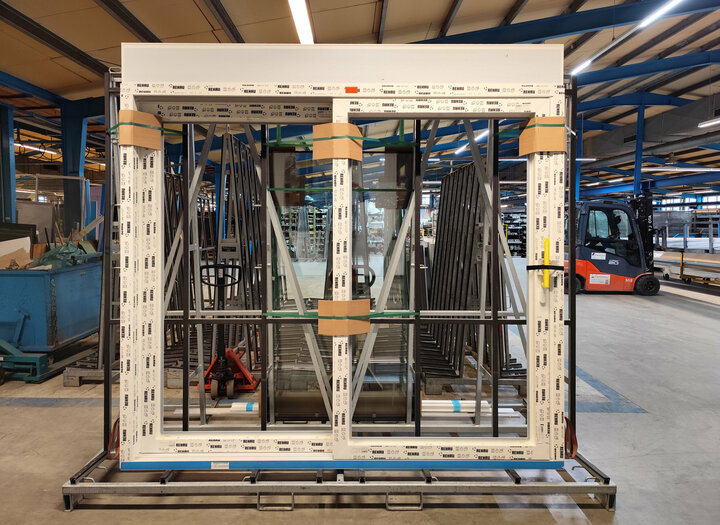 Ein großes Schiebe-Fenster  ist auf einem Metallgestell in einer Lagerhalle ausgestellt, begleitet von einem roten Gabelstapler und einem blauen Kran.