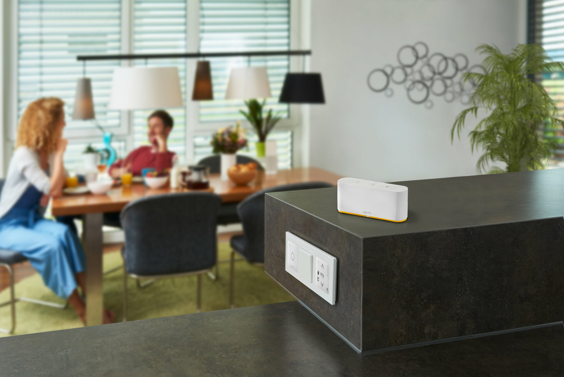 In einer modernen Küche stehen ein Esstisch aus Holz mit weißer Tischdecke, zwei Personen, die sich unterhalten, und ein weiß-orangefarbener Smart Speaker auf einer schwarzen Arbeitsplatte.