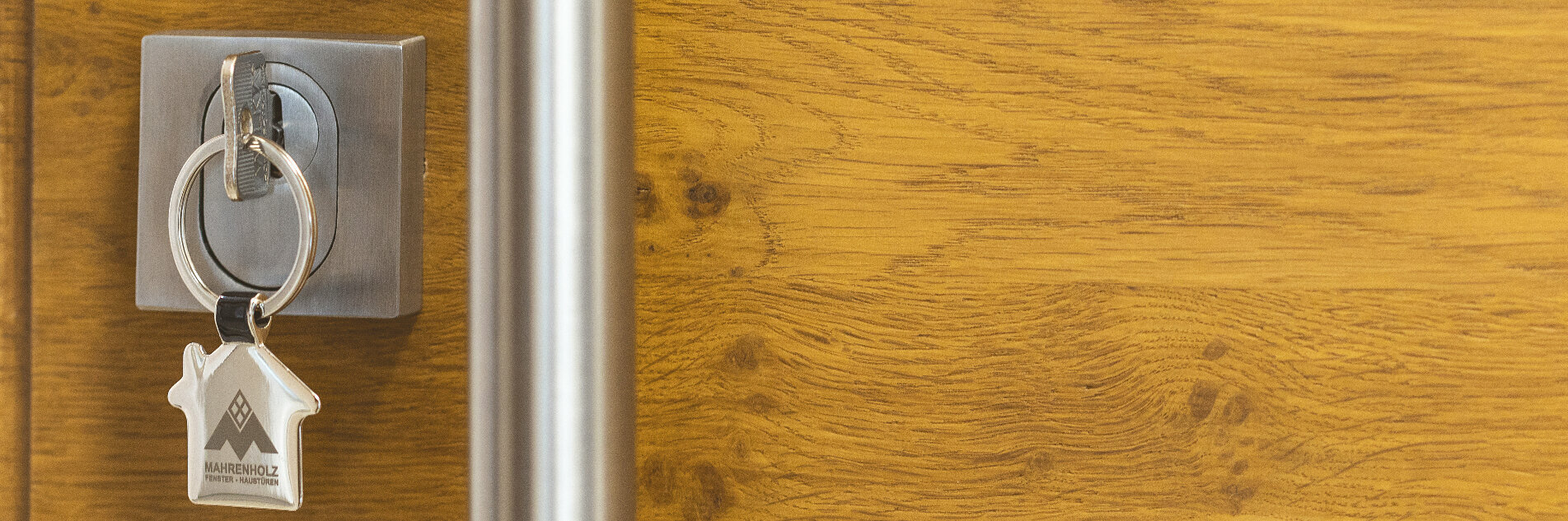 Ein silberner Schlüsselanhänger mit einem Hausschlüssel und dem Text &quot;MAHRENHOLZ&quot; hängt an einem silbernen Türgriff, der an einer Holztür mit Holzmaserung befestigt ist.