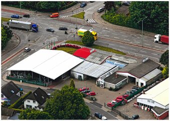 Eine Luftaufnahme einer belebten Stadtkreuzung zeigt ein großes weißes Gebäude mit rotem Dach, umgeben von kleineren Gebäuden, geparkten Autos und einer Mischung aus Fahrzeugen, mit einem gelben Lastwagen und einem roten Auto im Vordergrund.