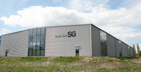 Ein modernes graues Gebäude mit einem großen Glasfenster und der Aufschrift "Stader Gas" steht auf einer grasbewachsenen Wiese, über der ein klarer blauer Himmel mit weißen Wolken hängt.