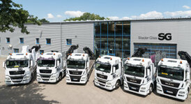 Sechs weiße Lastwagen mit schwarzen und roten Akzenten, darunter ein Kran, stehen vor einem grauen Gebäude mit einem blauen Glasfenster, auf dem das Logo "SG Stader Glas" prangt.