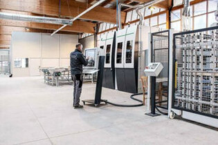 Ein Mann bedient eine große Industriemaschine in einer geräumigen Lagerhalle, umgeben von Maschinen und einer hohen Decke mit Holzbalken.