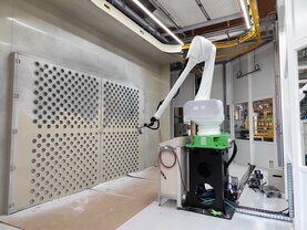 Ein weißer Roboterarm mit grünem und schwarzem Sockel, ausgestattet mit einer Spritzpistole, steht in einem sauberen, industriell anmutenden Raum mit einer Betonwand, gelben Rohren und einem Fenster.