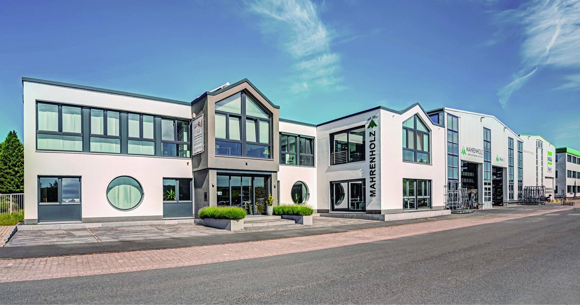 Ein modernes weißes Firmengebäude mit grauem Dach, großen Fenstern und einem Schild &quot;Mahrenholz&quot; steht an einer Straße, daran schließen eine Produktionshalle sowie weitere Gebäude an. Der Himmel ist blau mit einigen wenigen Wolken.