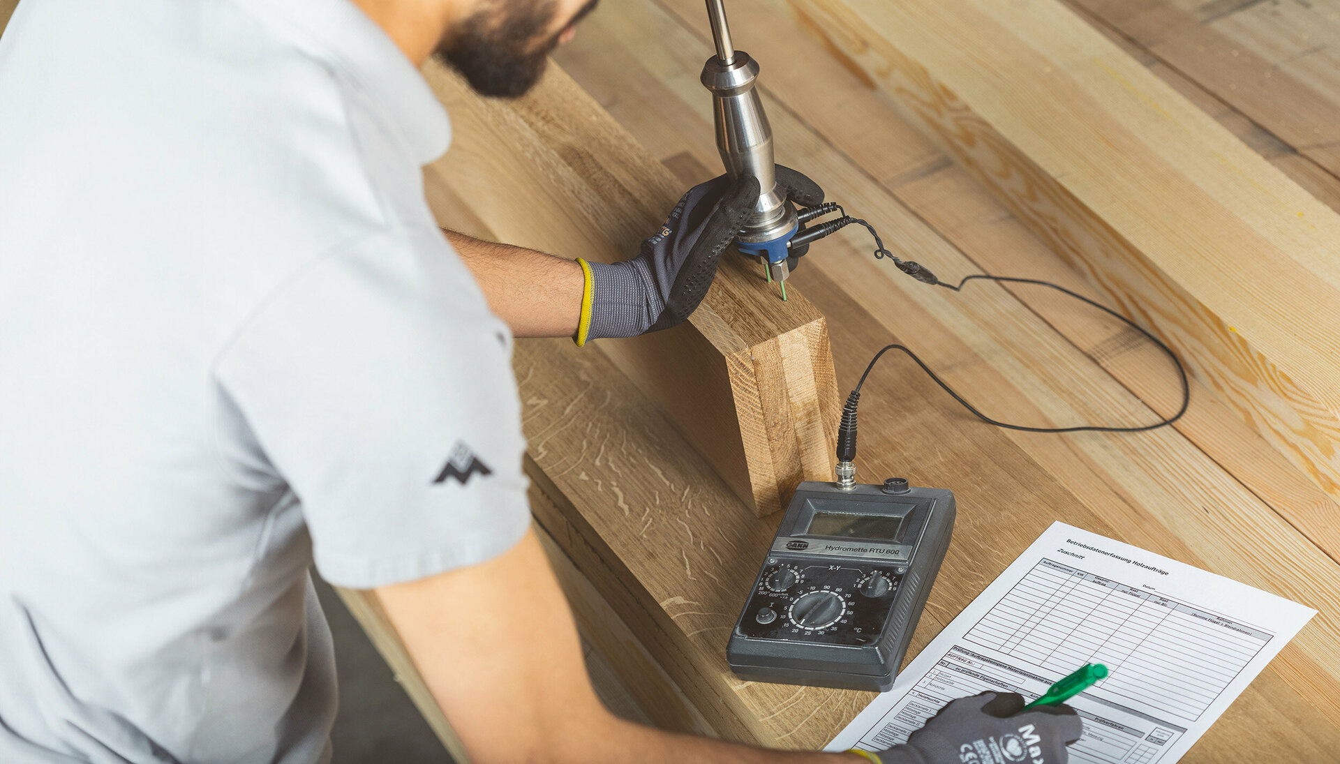 Eine Person in grauem Hemd und schwarzen Handschuhen prüft mit einem schwarz-silbernen Werkzeug ein Holzbrett, begleitet von einem grünen Stift und einem weißen Papier mit schwarzem Gitter.