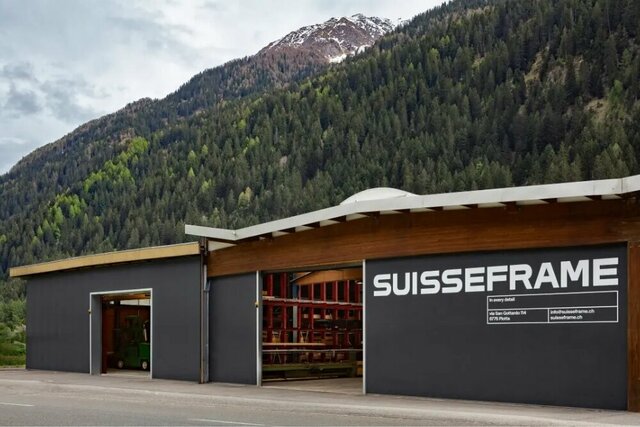Ein modernes Gebäude mit Holzfassade  steht an einer Straße in einer bergigen Gegend. Der Produktionsstandort SUISSE FRAME AG befindet sich im Tessin in der Schweiz.