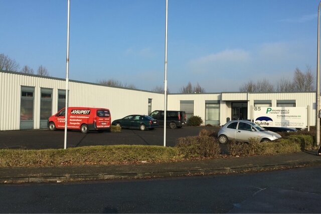 Ein weißes Gebäude, der Produktionsstandort in Lemgo, steht zentral an einem Parkplatz mit Autos. Der Himmel ist blau..