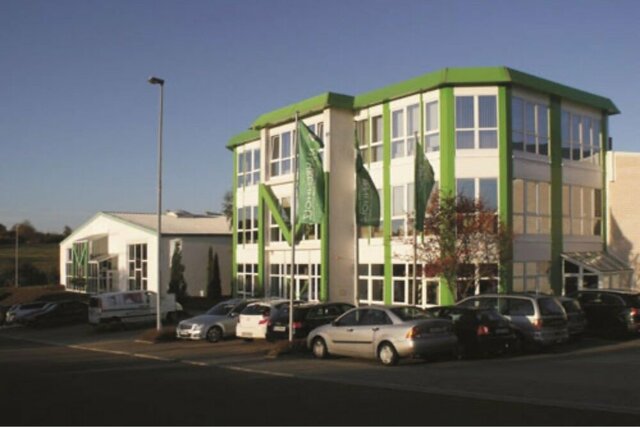 Ein modernes weißes und grünes Gebäude mit großen Fenstern und einem grünen Dach beherbergt ein Unternehmen, unsere Niederlassung in Jahnatal, umgeben von einem Parkplatz und einem klaren blauen Himmel.