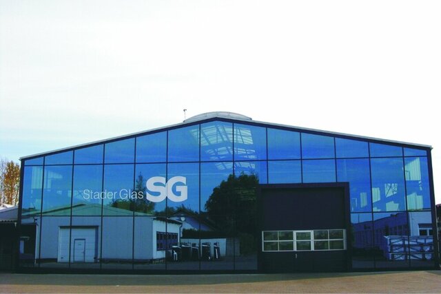 Ein modernes Glasgebäude mit großen Fenstern spiegelt einen Baum und ein weißes Gebäude. Ein Rolltor ist in der Fassade zu sehen sowie ein großes Logo "Stader Glas" auf der Glasfassade aufgedruckt..