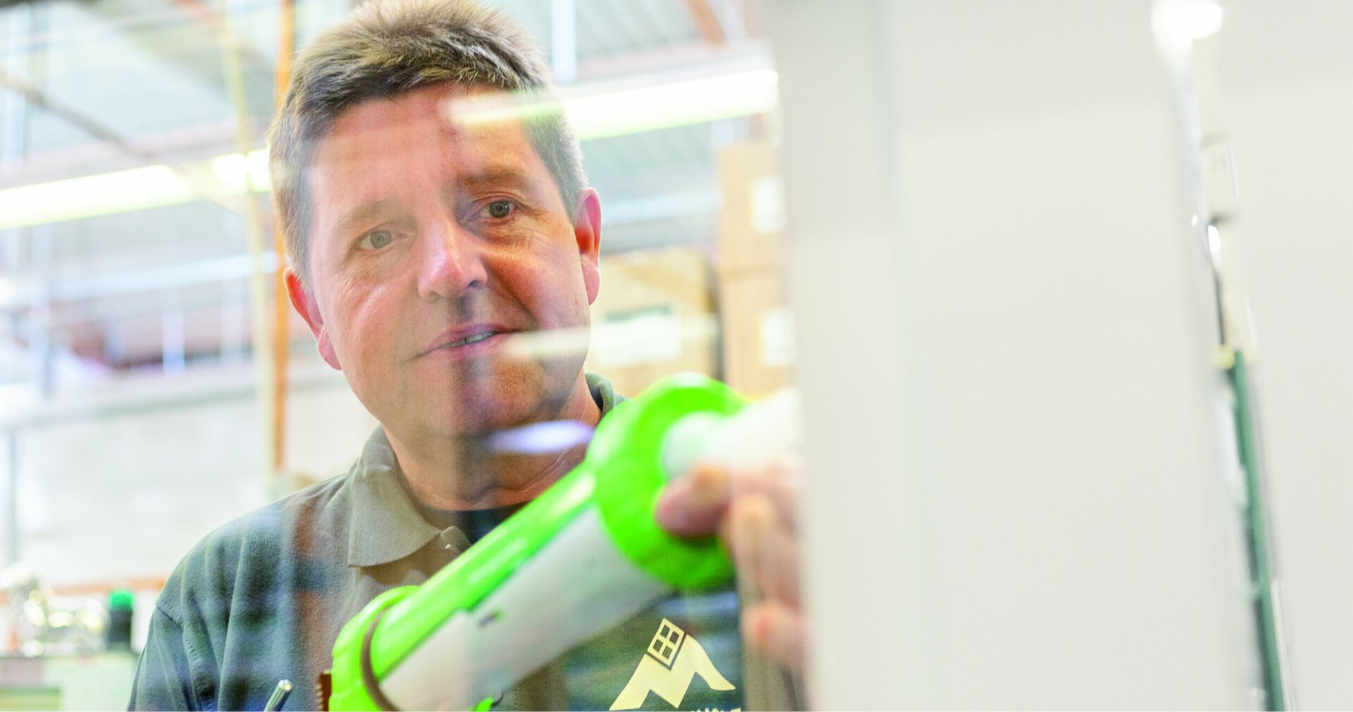Ein Mann in einem grauen Polohemd und mit einer grünen Spritzpistole in der Hand steht in einer Lagerhalle mit weißer Wand, versiegelt ein Fenster und blickt in die Kamera.