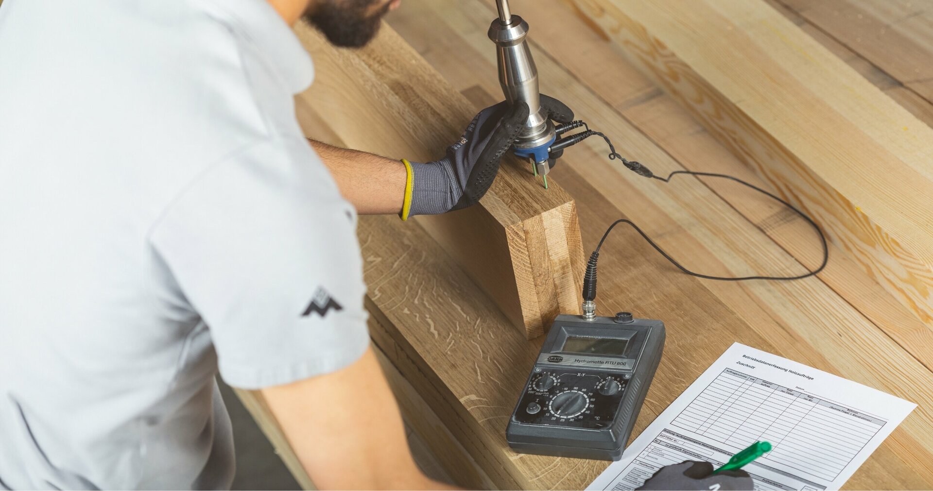 Eine Person in grauem Hemd und schwarzen Handschuhen prüft mit einer blau-silbernen Bohrmaschine die Festigkeit eines Holzbalkens, begleitet von einem schwarz-weißen Dokument und einem grünen Stift.