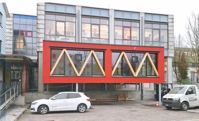 Ein modernes rot-weißes Gebäude mit großen Fenstern, vor dem Gebäude ist ein Parkplatz. Es handelt sich um einen neuen Showroom.